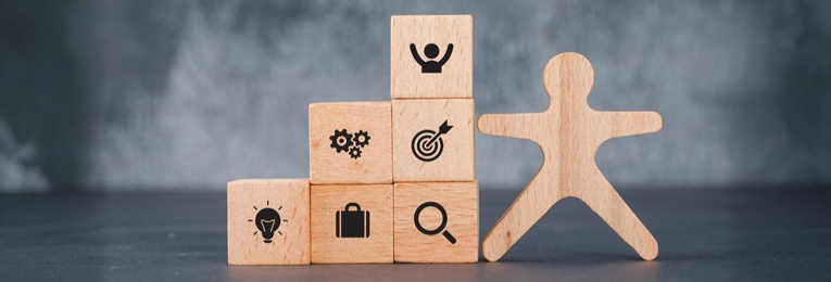 Six small wooden cubes and a wooden human-shaped figurine arranged on a grey table, with the cubes symbolizing diverse work goals through distinct markings or symbols.
